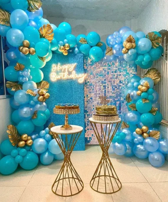 Shimmer backdrop birthday decoration with aqua blue, sky blue, turquoise, chrome gold, and metallic gold balloons, golden artificial leaves, cake table, and Happy Birthday wall sign in Dubai, UAE by Event Experts.