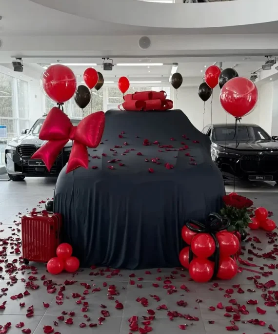 Car reveal balloon decoration setup with red balloons, big red foil bow, black red helium balloons, gift box balloons, white car cover and rose petals by Event Experts UAE