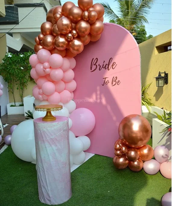 Bridal shower balloon arch setup with white, rose gold and pink balloons, white backdrop board, cake table, bride to be banner and floor balloon bunch by Event Experts UAE