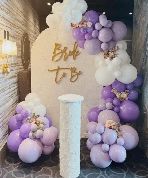 Bride balloon arch setup with light purple and dark purple balloons, white backdrop board and round cake table by Event Experts UAE