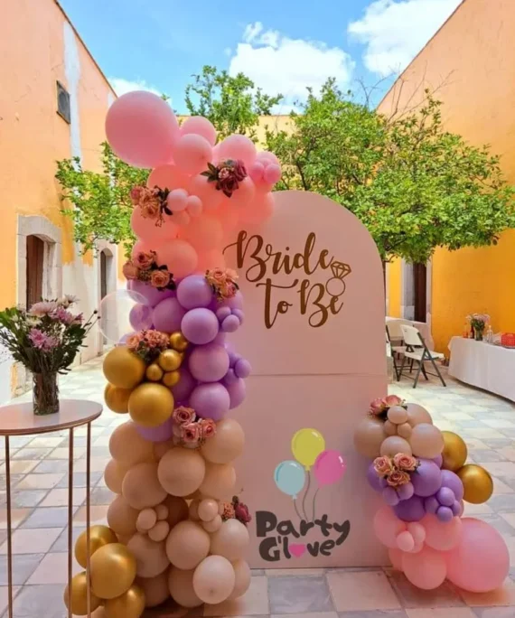 Bride welcome balloon arch setup with light pink, purple, golden and sand white balloons, white board backdrop, artificial flowers and golden cake stand by Event Experts UAE