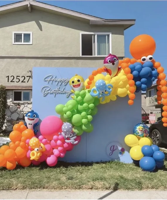 Baby shark theme balloon arch setup with blue yellow green pink orange balloons, 3 baby shark cutouts, light blue backdrop and happy birthday neon light by Event Experts UAE
