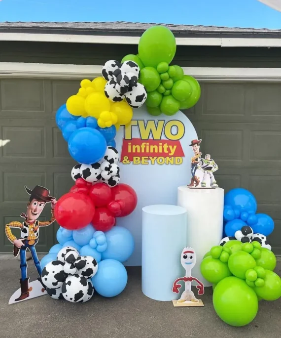 Toy Story theme kids balloon decoration setup with lime green, yellow, royal blue, light blue, red, and dark blue balloons, football color balloons, white round cake tables, and Toy Story character cutouts.