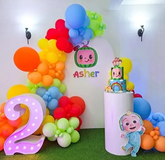 Cocomelon theme half arch balloon decoration setup with red, orange, yellow, green, and blue balloons, large balloons, white round cake table, Cocomelon cutout, and number cutout.