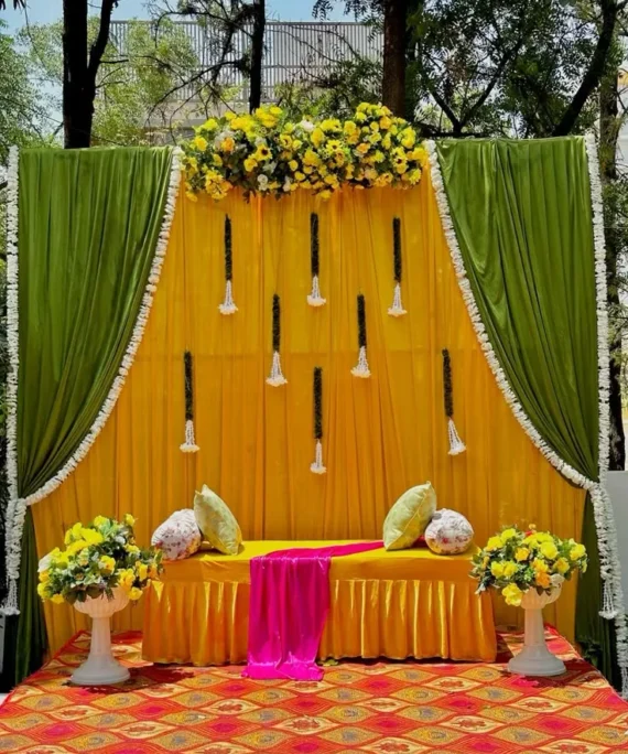 Mehndi curtain backdrop decoration setup with green and yellow curtains, artificial yellow, pink, and white flowers, sunflower arrangements, and large floral side bunches.