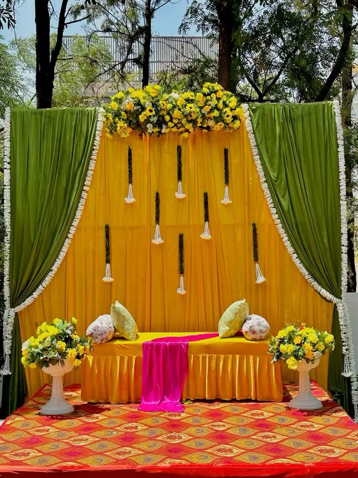 Mehndi curtain backdrop decoration setup with green and yellow curtains, artificial yellow, pink, and white flowers, sunflower arrangements, and large floral side bunches.