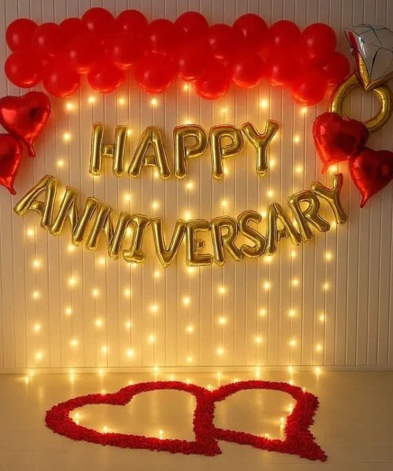 Simple Happy Anniversary balloon decoration setup with red balloons, red heart balloons, ring foil balloon, diamond foil balloon, Happy Anniversary banner, wall balloons, floor balloons, and red rose petals.