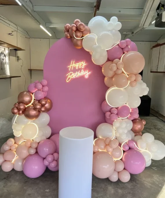 Half arch birthday balloon decoration setup with rose gold, peach, white, and bubblegum pink balloons, white round cake table, Happy Birthday neon light, and LED balloon lights.