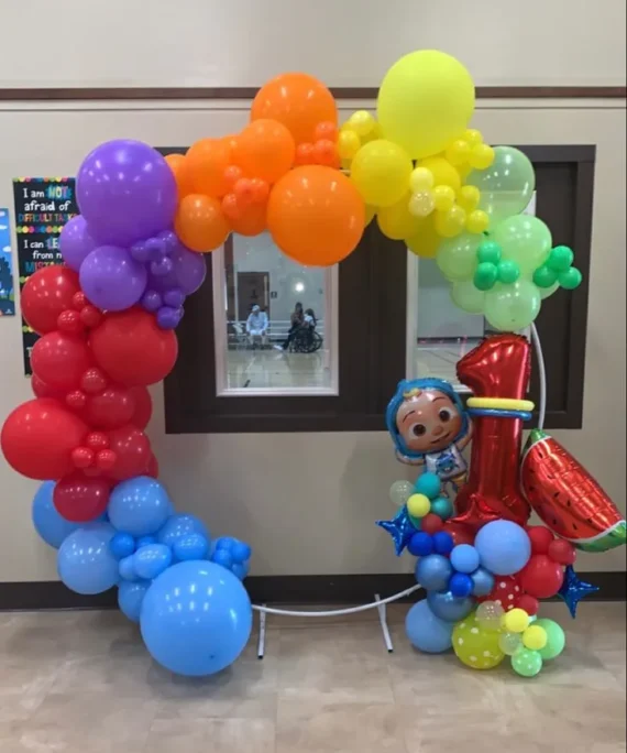 Cocomelon theme round arch balloon decoration setup with red, orange, yellow, green, light blue, and purple balloons, transparent balloons, number foil balloon, Cocomelon foil balloon, watermelon foil balloon, and white round cake table.