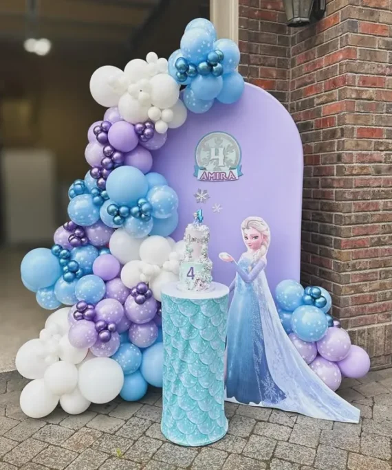 Frozen theme half arch backdrop balloon decoration with blue, purple, and white balloons, round white cake table, princess cutout, and personalized name print.