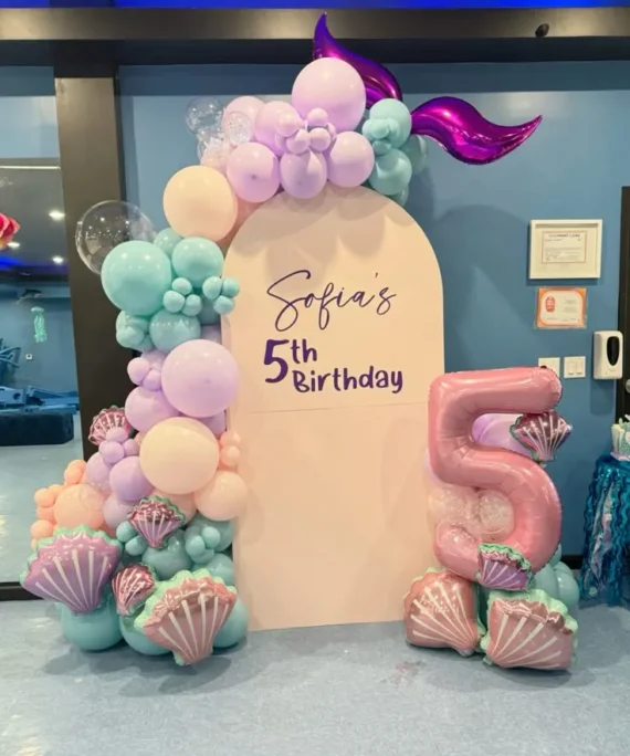 Mermaid theme half arch backdrop balloon decoration setup with light purple, pastel teal, soft pink, pink, transparent balloons, sea shell foil balloons, and light orange balloons.