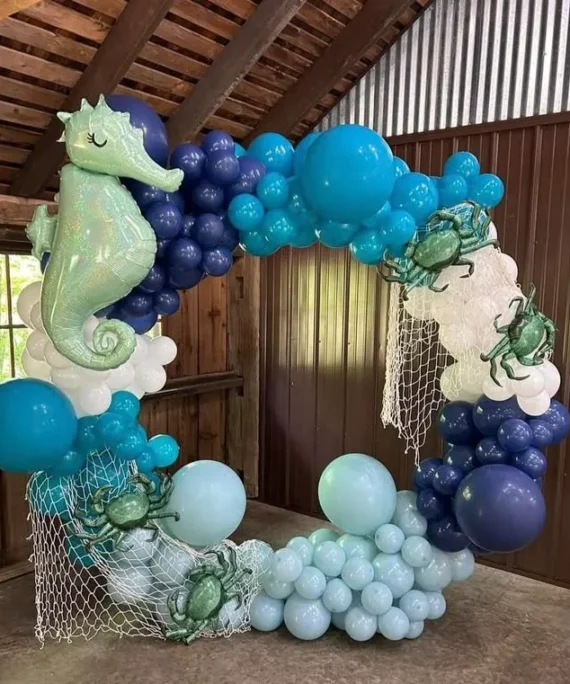 Mermaid theme round arch balloon decoration setup with navy blue, cyan, pastel blue, mint green, green, white balloons, one jumbo balloon, decorative fishing net, large seahorse foil balloon, and sea crab foil balloons.