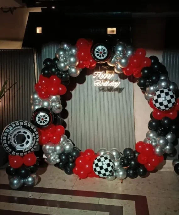 Kids car theme round arch balloon decoration setup with red, silver, and black balloons, disco balls, tire balloons, birthday neon light, and a medium round cake table.