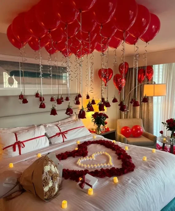 First night balloon decoration setup with red balloons, helium heart foil balloons, red rose petals, candle decoration, and red flower bouquet for romantic wedding room décor.
