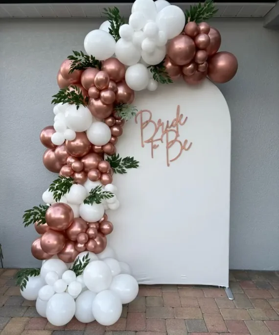 Bride To Be white cloth backdrop balloon decoration setup with rose gold and white balloons, artificial leaves, Bride To Be foil balloon, and round cake table.