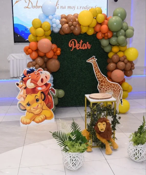 The Jungle Simba theme balloon backdrop setup with blue, orange, yellow, brown, and beige balloons, giraffe cutout, Simba character cutouts, artificial green leaves, and gold square metal cake table.