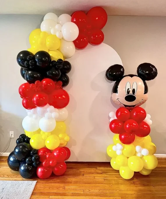 Mickey Mouse birthday backdrop balloon decoration setup with red, light yellow, white, and black balloons, Mickey Mouse foil balloon, and white round cake table.