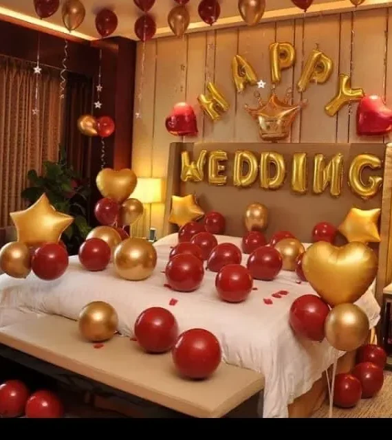 Happy Wedding balloon decoration setup with red and gold balloons, gold star foil balloons, red heart foil balloons, rose gold ring foil balloon, hanging balloons, rose petals, and romantic wedding room décor.