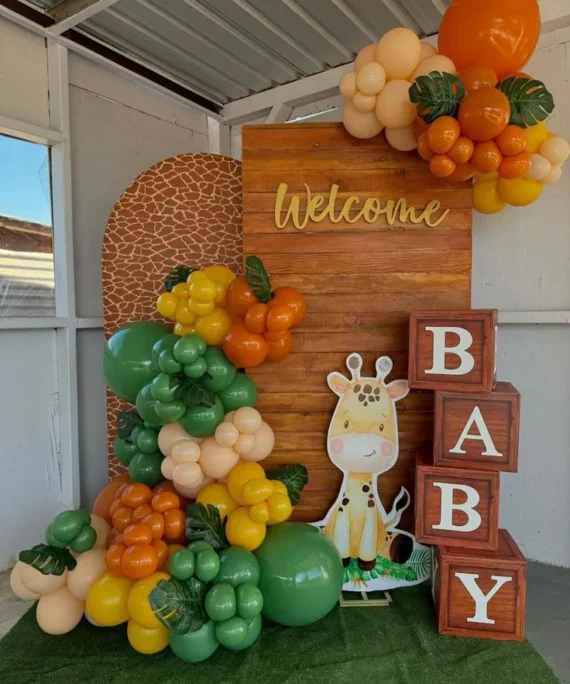 Jungle theme balloon decoration setup with green, orange, yellow, cream balloons, artificial green leaves, lion cutout, welcome banner, square frame backdrop, baby boxes, and green mat.