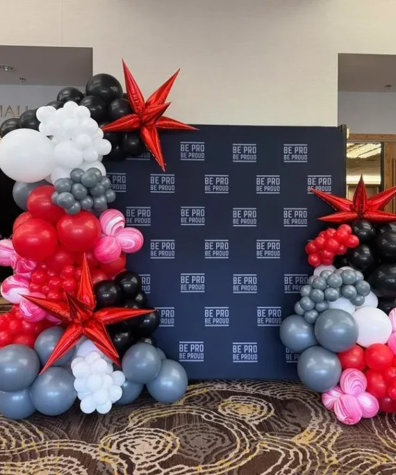 Corporate frame backdrop balloon decoration setup with black, white, gray, red, and pink balloons, large gray balloons, red star foil balloons, printed backdrop, and golden square cake tables.