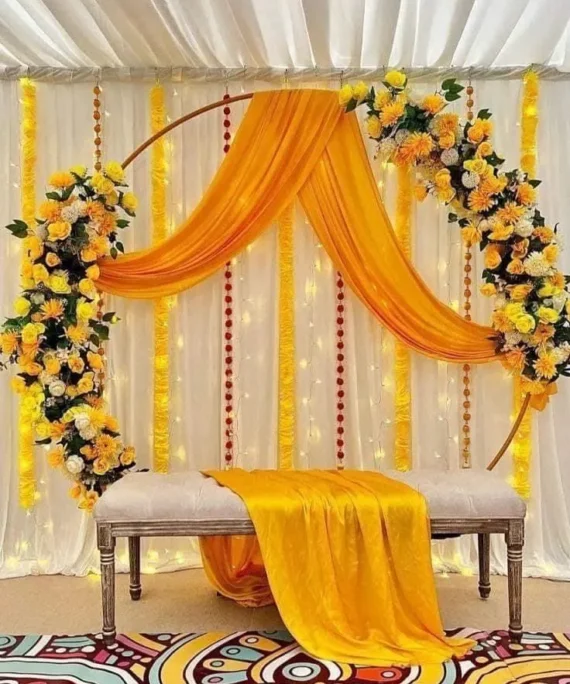 Haldi round arch frame flower decoration setup with yellow net fabric, white backdrop, artificial yellow, orange, and white flowers, artificial hay bales, LED lights, and yellow table net cloth.