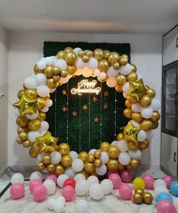 Anniversary round arch balloon decoration setup with gold, white, and pink balloons, gold foil stars, silver shimmer strands, Happy Anniversary foil banner, ceiling balloons, and floor balloons.