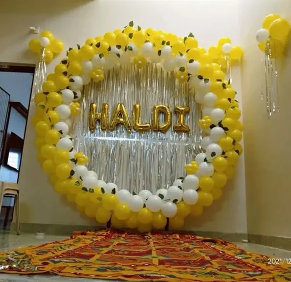 Haldi round arch balloon decoration setup with yellow and white balloons, golden Haldi foil banner, silver shimmer backdrop, green artificial leaves, wall balloon bunches, and floor balloons.