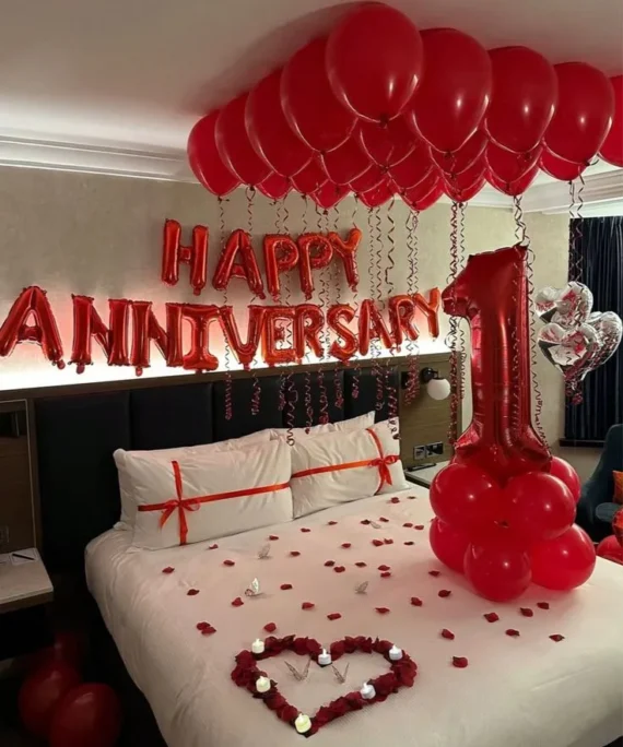 1st anniversary balloon decoration setup with red balloons, Happy Anniversary foil banner, number foil balloon, ceiling balloons with ribbons, silver heart helium balloons, flower heart, silver butterflies, rose petals, and LED candles.