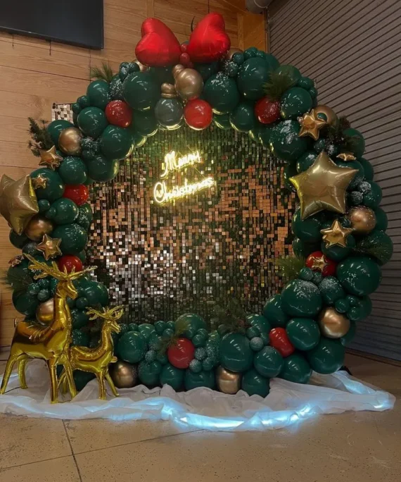 Merry Christmas round frame silver shimmer backdrop with green and red balloons, golden star foil balloons, green garlands, golden deer, LED lights, white cloth, snow décor, and red heart foil balloons.